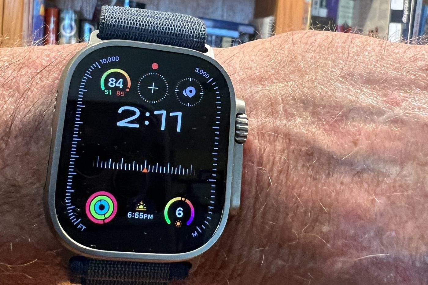 An Apple Watch Ultra 2 with a grey woven band, worn on an older man’s wrist. The face shows the time 2:11, a heart rate of 84, and coloured activity rings. Bookshelves fill the background.